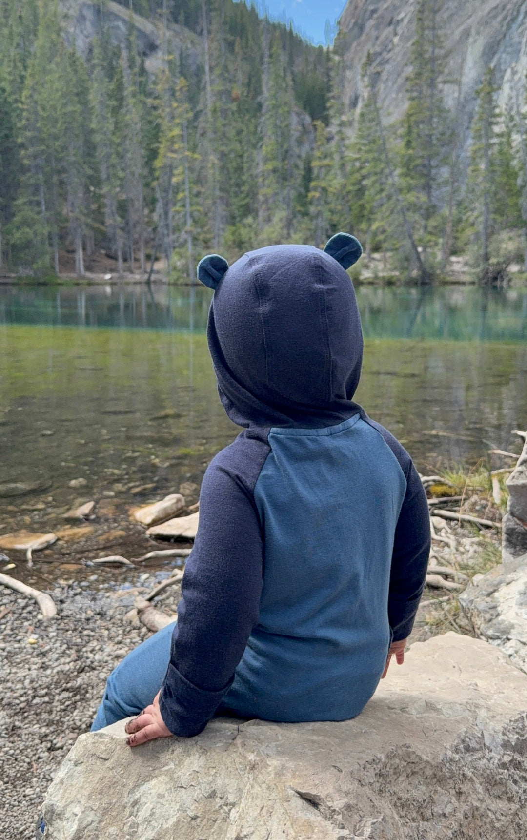Child in bear-themed hoodie sitting by a lake with mountains in the background